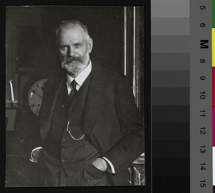 Portrait en pied du Prof. Edouard de Cérenville, appuyé contre une cheminée en marbre. Derrière lui, un miroir. A sa gauche, des bougies et une horloge, des photographies. A sa droite des tableaux sont accrochés au mur.